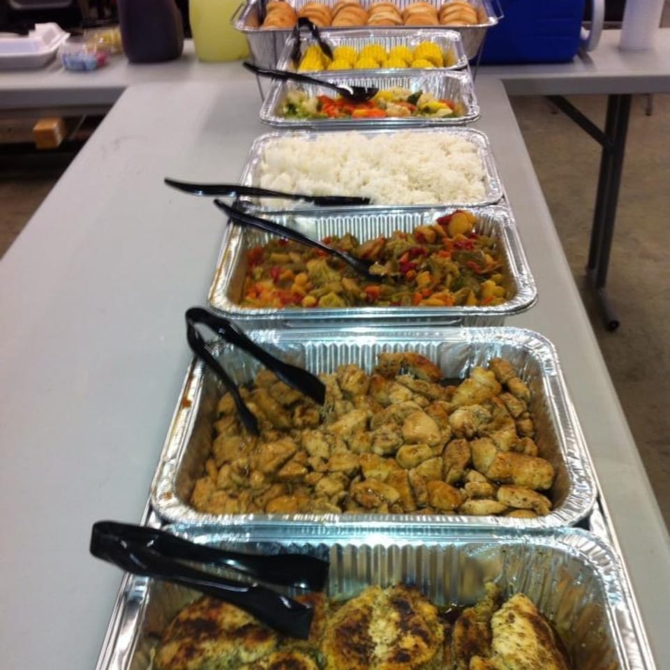 Catering trays of rice, vegetables, chicken, and rolls on a table.