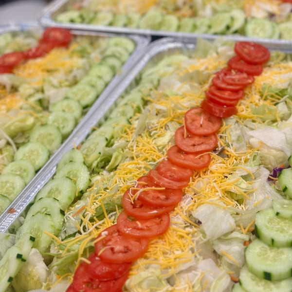 Multiple catering trays of fresh garden salad with lettuce, shredded cheese, sliced tomatoes, and cucumbers.